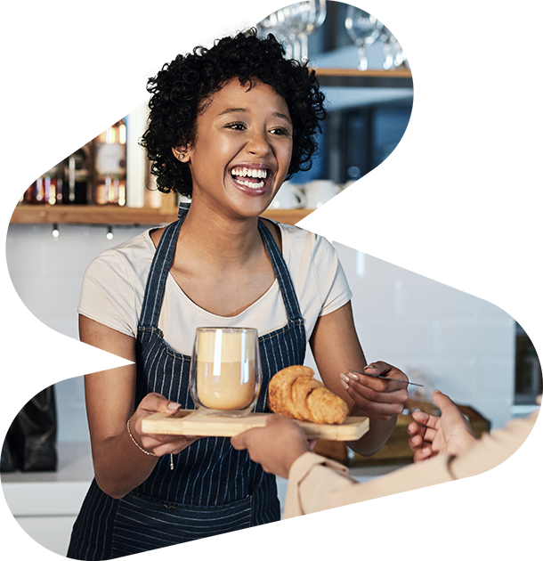 Smiling woman in an apron serving a tray with a coffee and croissants, handing it to a customer in a cozy café setting.